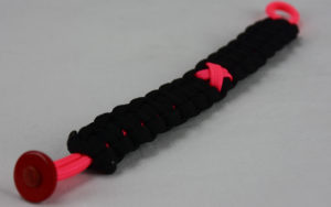 pink and black breast cancer support paracord bracelet with pink ribbon and red button fastener in the bottom corner